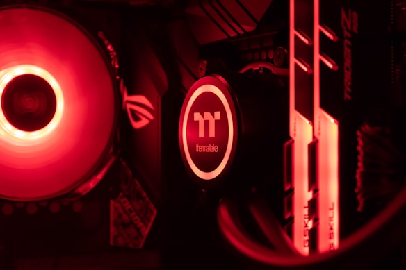 A close-up view of a computer's internal components, featuring a glowing red cooling fan, a Thermaltake CPU cooler with illuminated branding, and two illuminated RAM sticks. The scene is dominated by a red lighting effect, enhancing the high-tech aesthetic.