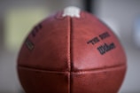 A close-up view of a leather football with textured surface and visible stitching. The ball is branded with text, including the words 'The Duke' and 'Wilson', printed in black ink.