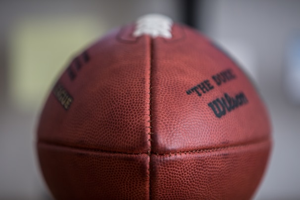 A close-up view of a leather football with textured surface and visible stitching. The ball is branded with text, including the words 'The Duke' and 'Wilson', printed in black ink.