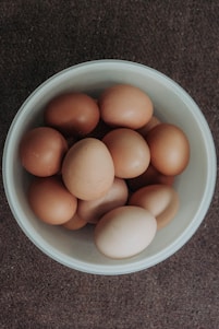 A white bowl filled with multiple brown eggs sits on a textured brown surface. The eggs vary slightly in shade, with some being darker than others.