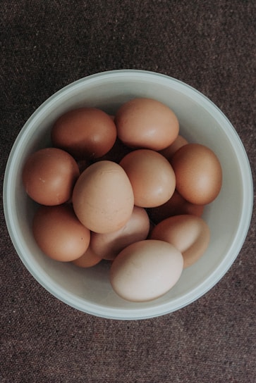 A white bowl filled with multiple brown eggs sits on a textured brown surface. The eggs vary slightly in shade, with some being darker than others.