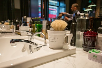 A bathroom or barbershop sink with a sleek chrome faucet is seen in a brightly lit environment. A white mug labeled 'Ted Baker London' containing a shaving brush sits on the countertop. Various bottles and grooming products are lined up in the background. A person is blurred in the background, suggesting movement or focus on a task.