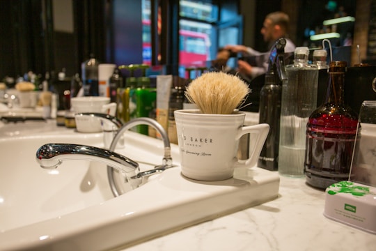 A bathroom or barbershop sink with a sleek chrome faucet is seen in a brightly lit environment. A white mug labeled 'Ted Baker London' containing a shaving brush sits on the countertop. Various bottles and grooming products are lined up in the background. A person is blurred in the background, suggesting movement or focus on a task.