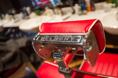 A close-up of a red barber chair with the name 'Belmont' prominently displayed on its shiny metallic headrest. The background features vague, out-of-focus shapes that seem to be other barber chairs and tools in the salon.