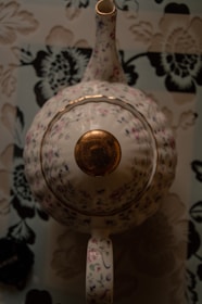 A floral-patterned teapot with a glossy, gold-colored lid sits on a matching floral background. The teapot's spout, lid, and handle are prominently shown.