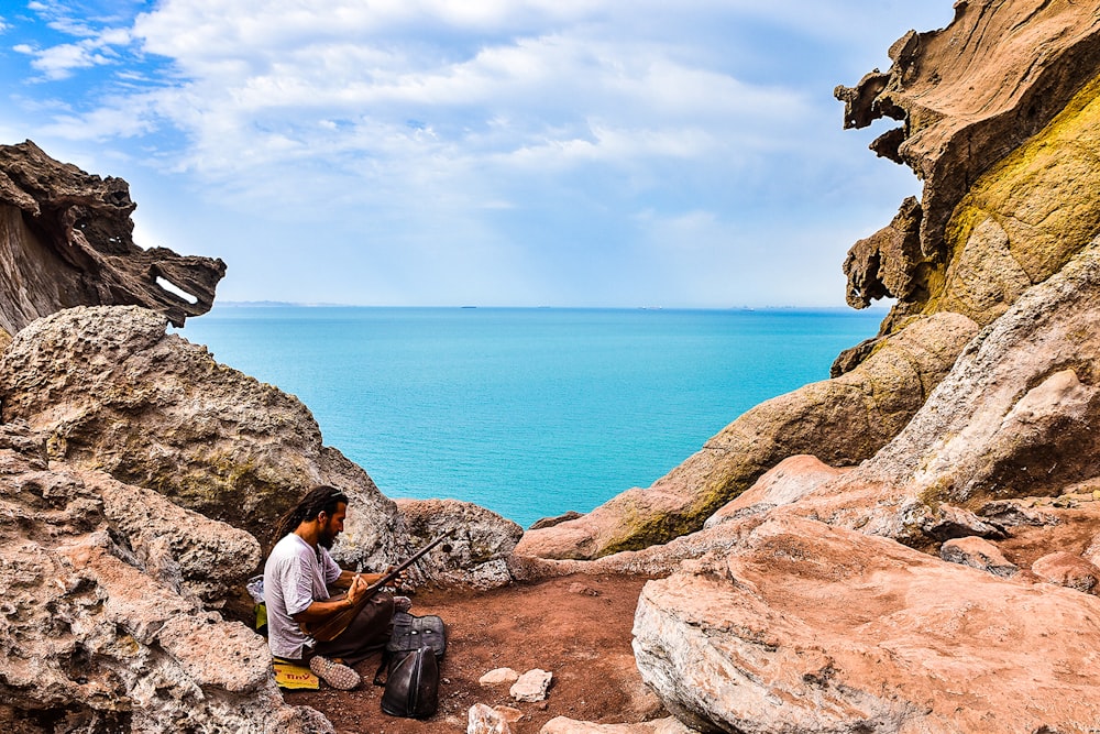 homem na jaqueta laranja sentado na formação rochosa marrom perto do corpo de água durante o dia