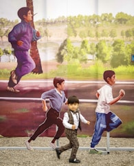 A young child dressed in formal attire is running playfully along an outdoor artwork featuring life-sized images of children running in the background. The mural depicts a lively and natural setting with trees and greenery.