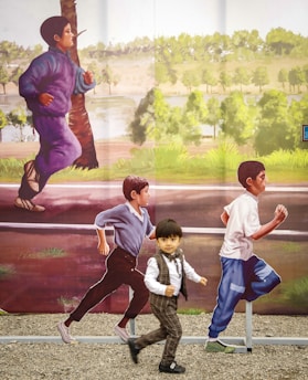 A young child dressed in formal attire is running playfully along an outdoor artwork featuring life-sized images of children running in the background. The mural depicts a lively and natural setting with trees and greenery.