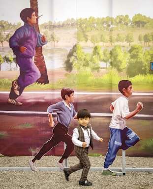 A young child dressed in formal attire is running playfully along an outdoor artwork featuring life-sized images of children running in the background. The mural depicts a lively and natural setting with trees and greenery.