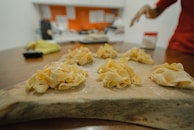 A wooden surface holds several nests of homemade pasta, dusted with flour. In the background, a blurred kitchen setting is visible with hints of red and white colors, and an out-of-focus hand appears on the right side of the image.
