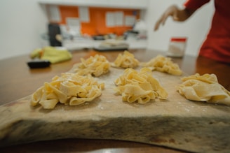 A wooden surface holds several nests of homemade pasta, dusted with flour. In the background, a blurred kitchen setting is visible with hints of red and white colors, and an out-of-focus hand appears on the right side of the image.