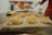 A wooden surface holds several nests of homemade pasta, dusted with flour. In the background, a blurred kitchen setting is visible with hints of red and white colors, and an out-of-focus hand appears on the right side of the image.