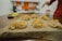 A wooden surface holds several nests of homemade pasta, dusted with flour. In the background, a blurred kitchen setting is visible with hints of red and white colors, and an out-of-focus hand appears on the right side of the image.