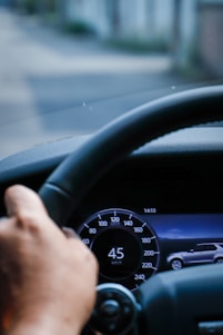 A close-up view of a car dashboard showing a digital speedometer reading 45 km/h. The image also includes a hand on the steering wheel and a screen displaying a vehicle graphic, with the time shown as 14:13. The background appears slightly blurred, indicating movement.