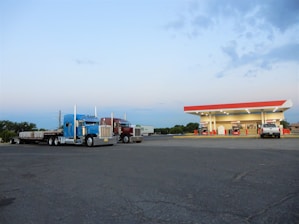 Two large semi-trucks are parked in a spacious asphalt lot, adjacent to a gas station with a red and white canopy. The gas station has several fuel pumps and is illuminated by bright lights. A smaller white vehicle is parked near the station. The sky is partly cloudy and the setting appears to be in an open, semi-rural area.