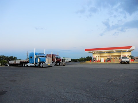 Two large semi-trucks are parked in a spacious asphalt lot, adjacent to a gas station with a red and white canopy. The gas station has several fuel pumps and is illuminated by bright lights. A smaller white vehicle is parked near the station. The sky is partly cloudy and the setting appears to be in an open, semi-rural area.