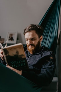 A man with a beard is comfortably seated in a hammock, reading a book titled 'Wild at Heart'. The lighting is soft, creating a relaxed atmosphere. In the background, there are blurred framed pictures and a light-colored wall.