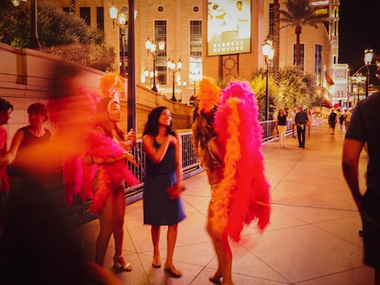 A vibrant street scene at night with people interacting. Two performers wearing colorful feathered costumes, including headpieces, are talking to a woman in a blue dress. The background features illuminated buildings, palm trees, and decorative street lamps, contributing to a lively and festive atmosphere.