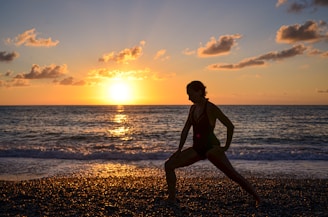 A person is performing a stretching exercise on a pebbled beach at sunset. The sky is filled with scattered clouds, and the sun is low on the horizon, casting a warm glow over the scene. The sea is calm with gentle waves reaching the shore.