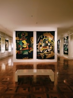 An art gallery room featuring two large colorful abstract paintings hanging side by side on a wall. The floor is made of polished wood, reflecting the artwork. A simple white bench is positioned in the center of the room, inviting viewers to sit and observe the art. There are additional paintings visible along the walls, under soft lighting.
