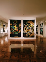 An art gallery room featuring two large colorful abstract paintings hanging side by side on a wall. The floor is made of polished wood, reflecting the artwork. A simple white bench is positioned in the center of the room, inviting viewers to sit and observe the art. There are additional paintings visible along the walls, under soft lighting.