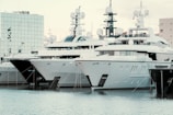 Several large yachts are docked in a harbor, with modern, angular designs. The boats are closely packed in a row, and their white and dark grey hulls reflect on the water. There is a modern building with mirrored glass in the background, adding to the contemporary feel of the scene.