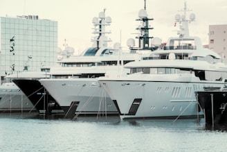 Several large yachts are docked in a harbor, with modern, angular designs. The boats are closely packed in a row, and their white and dark grey hulls reflect on the water. There is a modern building with mirrored glass in the background, adding to the contemporary feel of the scene.
