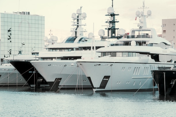 Several large yachts are docked in a harbor, with modern, angular designs. The boats are closely packed in a row, and their white and dark grey hulls reflect on the water. There is a modern building with mirrored glass in the background, adding to the contemporary feel of the scene.