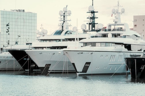 Several large yachts are docked in a harbor, with modern, angular designs. The boats are closely packed in a row, and their white and dark grey hulls reflect on the water. There is a modern building with mirrored glass in the background, adding to the contemporary feel of the scene.