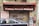 A closed storefront with the sign L'Ingenieur above, offering books, gifts, and toys. The metal shutter is covered in graffiti, and there is a prominent maroon-colored awning. A parked car half-blocks the view in front, with additional signs on the side listing office supplies, drawing materials, and photocopy services.