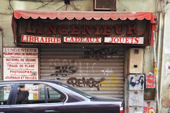 A closed storefront with the sign L'Ingenieur above, offering books, gifts, and toys. The metal shutter is covered in graffiti, and there is a prominent maroon-colored awning. A parked car half-blocks the view in front, with additional signs on the side listing office supplies, drawing materials, and photocopy services.