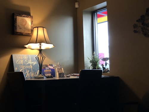 A cozy interior scene features a table with a lamp casting a warm glow. On the table, there is a decorative sign with a motivational message, and a plant is visible next to some books and a framed picture. A window allows natural light to enter, complementing the lamp's illumination.