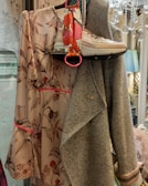 A floral patterned dress with shades of pink and brown is displayed alongside a pair of beige high-top sneakers. A herringbone wool coat, predominantly grey, is draped next to the dress. There is a colorful scarf with bright red and pink hues near the sneakers, adding a pop of color. A decorative chandelier hangs in the upper right corner, contributing to an elegant setting.