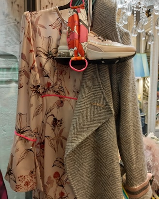 A floral patterned dress with shades of pink and brown is displayed alongside a pair of beige high-top sneakers. A herringbone wool coat, predominantly grey, is draped next to the dress. There is a colorful scarf with bright red and pink hues near the sneakers, adding a pop of color. A decorative chandelier hangs in the upper right corner, contributing to an elegant setting.