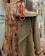 A floral patterned dress with shades of pink and brown is displayed alongside a pair of beige high-top sneakers. A herringbone wool coat, predominantly grey, is draped next to the dress. There is a colorful scarf with bright red and pink hues near the sneakers, adding a pop of color. A decorative chandelier hangs in the upper right corner, contributing to an elegant setting.