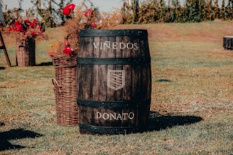 A wooden barrel with the words 'Viñedos Donato' engraved on it sits on a grassy field. To the left of the barrel, there are wicker baskets filled with colorful flowers including red roses.