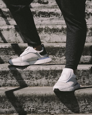 A person wearing white sneakers is walking up concrete steps. The sneakers have a modern design with noticeable cushioning and a textured surface. The steps appear to be weathered, with visible discoloration and subtle shadows casting across them. The outfit is primarily dark pants, providing contrast with the bright footwear.