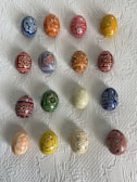 Fifteen intricately decorated eggs are arranged in a neat grid on a textured white surface. Each egg features unique patterns and colors, with designs ranging from floral to geometric motifs. The collection displays a diversity of colors, including blues, reds, oranges, and yellows.