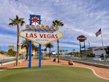 A famous sign welcomes visitors with the text 'Welcome to Fabulous Las Vegas, Nevada' in bright colors. Surrounded by palm trees, the scene features a clear blue sky with scattered clouds. In the background, iconic Las Vegas architecture and an American flag are visible, contributing to a lively and iconic city landscape.