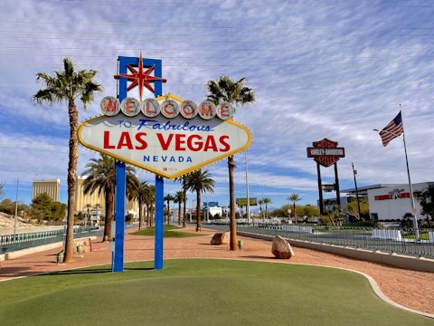 A famous sign welcomes visitors with the text 'Welcome to Fabulous Las Vegas, Nevada' in bright colors. Surrounded by palm trees, the scene features a clear blue sky with scattered clouds. In the background, iconic Las Vegas architecture and an American flag are visible, contributing to a lively and iconic city landscape.