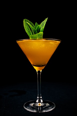A tall martini glass containing a vibrant orange cocktail, garnished with a cluster of fresh mint leaves, set against a stark black background.