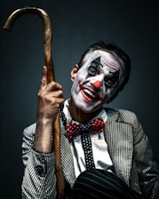 A person dressed as a clown with face makeup featuring white, black, and red colors. The individual is smiling broadly and holding a wooden cane. The outfit includes a pinstriped suit and a red polka-dot bow tie, creating a playful yet somewhat eerie appearance.