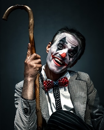 A person dressed as a clown with face makeup featuring white, black, and red colors. The individual is smiling broadly and holding a wooden cane. The outfit includes a pinstriped suit and a red polka-dot bow tie, creating a playful yet somewhat eerie appearance.