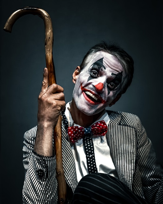 A person dressed as a clown with face makeup featuring white, black, and red colors. The individual is smiling broadly and holding a wooden cane. The outfit includes a pinstriped suit and a red polka-dot bow tie, creating a playful yet somewhat eerie appearance.