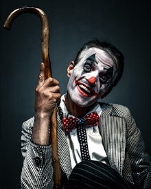 A person dressed as a clown with face makeup featuring white, black, and red colors. The individual is smiling broadly and holding a wooden cane. The outfit includes a pinstriped suit and a red polka-dot bow tie, creating a playful yet somewhat eerie appearance.