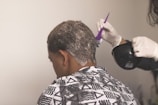 A person is sitting with their back to the camera, receiving a hair treatment. A second individual wearing gloves is applying a white substance to the person's hair using a purple brush, holding a black bowl in their other hand. The person receiving the treatment is draped in a patterned cape featuring triangles and lines.