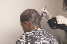 A person is sitting with their back to the camera, receiving a hair treatment. A second individual wearing gloves is applying a white substance to the person's hair using a purple brush, holding a black bowl in their other hand. The person receiving the treatment is draped in a patterned cape featuring triangles and lines.