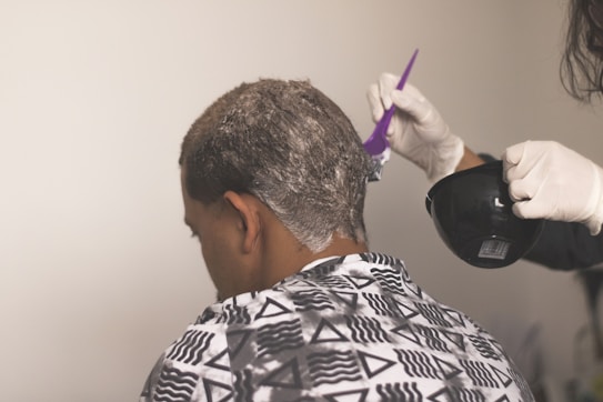 A person is sitting with their back to the camera, receiving a hair treatment. A second individual wearing gloves is applying a white substance to the person's hair using a purple brush, holding a black bowl in their other hand. The person receiving the treatment is draped in a patterned cape featuring triangles and lines.