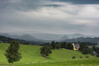 A serene landscape featuring a lush green field with scattered trees in the foreground, and a quaint church with a steeple nestled among more trees in the mid-ground. The background showcases rolling hills fading into the horizon under an expansive, overcast sky.