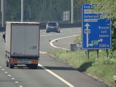 A highway scene with a truck driving on the road. A large blue road sign shows directions to Gent, Antwerpen, Brussel, Luik, and Leuven via the E40 motorway. There are clear lane markings and a car in the distance. Green trees and grass line the roadside.